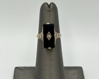 May include: A silver-toned ring featuring a rectangular black onyx stone set in the center. A small, clear, diamond-shaped accent stone is embedded in the middle of the onyx. The band displays intricate, openwork filigree metal detailing.