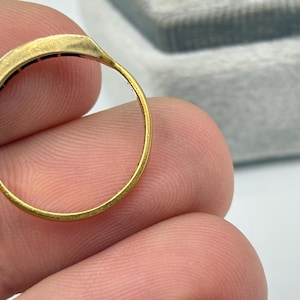 May include: Close-up of a gold ring with a rectangular top. The ring has a thin band and a row of small, dark stones set into the top. The ring is held between fingers.