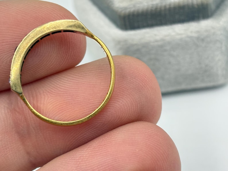 May include: Close-up of a gold ring with a rectangular top. The ring has a thin band and a row of small, dark stones set into the top. The ring is held between fingers.