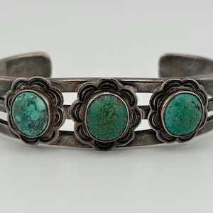 May include: A silver cuff bracelet featuring three round turquoise stones set in scalloped bezels. The band is constructed from three parallel silver wires that connect at the ends, showcasing a dark, oxidized finish on the metal surface.
