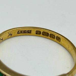 May include: Close-up of a gold ring with a band featuring small, colorful gemstones. The ring has markings including "N", "1/2", and "ACCS". The gemstones appear to be green and dark blue. The ring is likely vintage or antique.