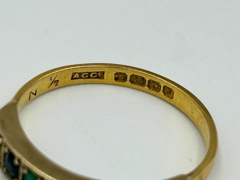 May include: Close-up of a gold ring with a band featuring small, colorful gemstones. The ring has markings including "N", "1/2", and "ACCS". The gemstones appear to be green and dark blue. The ring is likely vintage or antique.