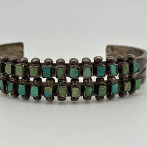 May include: A silver cuff bracelet featuring two parallel rows of small, rectangular, turquoise stones set in metal bezels. The design includes decorative silver beads between each stone, and the band has a subtle etched pattern on the sides.
