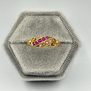 May include: A gold ring featuring a curved, wave-like design set with a row of vibrant pink rubies and a parallel row of small, round white pearls. The band has decorative scrollwork details at both ends of the central gemstone setting.