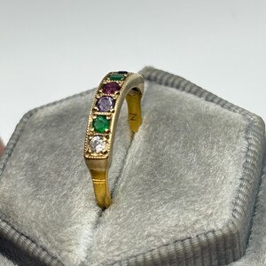 May include: A gold ring with a row of colorful gemstones, including green, red, purple, and white stones. The ring is set against a gray velvet background, highlighting the vibrant colors of the gemstones. The ring's design is elegant and eye-catching.