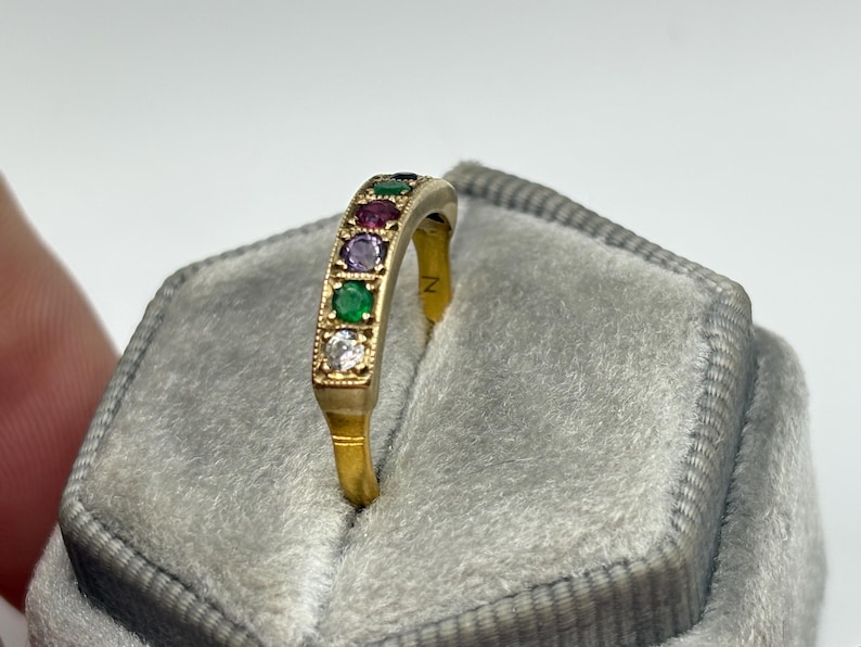 May include: A gold ring with a row of colorful gemstones, including green, red, purple, and white stones. The ring is set against a gray velvet background, highlighting the vibrant colors of the gemstones. The ring's design is elegant and eye-catching.