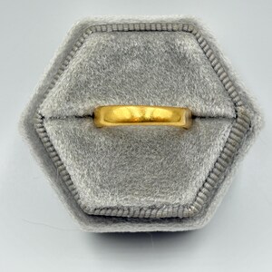 May include: A gold ring with a flat band rests on a gray velvet ring box. The ring has a polished, shiny appearance. The ring box is hexagonal in shape and has a soft, textured surface.