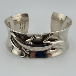 May include: A wide sterling silver cuff bracelet featuring a raised, three-dimensional lizard design across the front. The interior of the cuff is stamped with the text "STERLING" and "MARY-RITA PADILLA" in a clear, legible font.