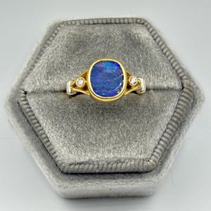 May include: A gold ring with a large, oval, blue opal center stone. The ring has two small diamonds on either side of the opal. The ring is set on a gray velvet surface.