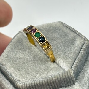 May include: A gold ring with a row of colorful gemstones, including red, green, and blue stones. The ring is set on a gray velvet surface, showcasing the ring's design and colors. The ring's band is a simple, elegant design.