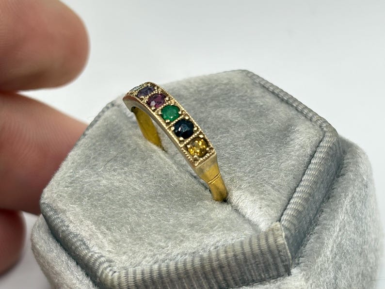 May include: A gold ring with a row of colorful gemstones, including red, green, and blue stones. The ring is set on a gray velvet surface, showcasing the ring's design and colors. The ring's band is a simple, elegant design.
