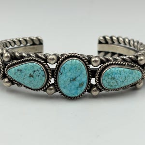 May include: A silver cuff bracelet featuring five turquoise stones in varying shapes, including a central oval and four teardrop-shaped stones. The metal band is decorated with twisted rope detailing and small silver spheres between the stones.