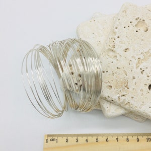 May include: A stack of silver wire bracelets, each about 6 cm in diameter, arranged in a circular pattern. The bracelets are on a white surface with a wooden ruler for scale.