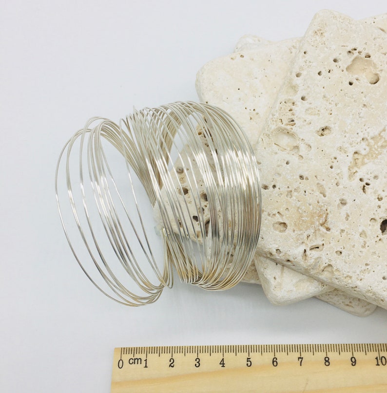May include: A stack of silver wire bracelets, each about 6 cm in diameter, arranged in a circular pattern. The bracelets are on a white surface with a wooden ruler for scale.