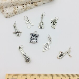 Peut inclure: Un ensemble de huit breloques d'instruments de musique argent&eacute;s, comprenant un violon, un saxophone, un microphone, une batterie, une cl&eacute; de sol, une cl&eacute; de fa, une trompette et une note de musique. Les breloques sont dispos&eacute;es sur une surface blanche avec une r&egrave;gle en bois pour l'&eacute;chelle.