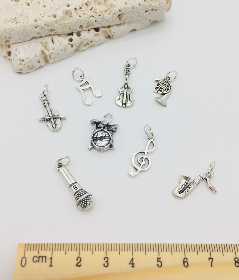 Peut inclure: Un ensemble de huit breloques d'instruments de musique argent&eacute;s, comprenant un violon, un saxophone, un microphone, une batterie, une cl&eacute; de sol, une cl&eacute; de fa, une trompette et une note de musique. Les breloques sont dispos&eacute;es sur une surface blanche avec une r&egrave;gle en bois pour l'&eacute;chelle.