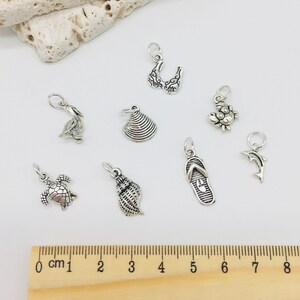 Peut inclure: Un ensemble de neuf breloques en argent avec diff&eacute;rents motifs sur le th&egrave;me de la plage, notamment un coquillage, un crabe, un dauphin, une tortue, une tong, un coquillage, un oiseau et un haut de bikini. Les breloques sont dispos&eacute;es sur une surface blanche avec une r&egrave;gle pour l'&eacute;chelle.