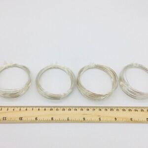 May include: Four spools of silver wire, each wound into a circle, are arranged in a row on a white surface. A wooden ruler with centimeter markings is below the spools.