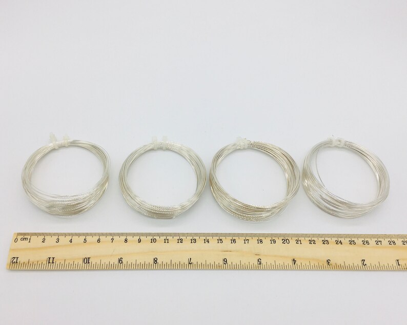 May include: Four spools of silver wire, each wound into a circle, are arranged in a row on a white surface. A wooden ruler with centimeter markings is below the spools.