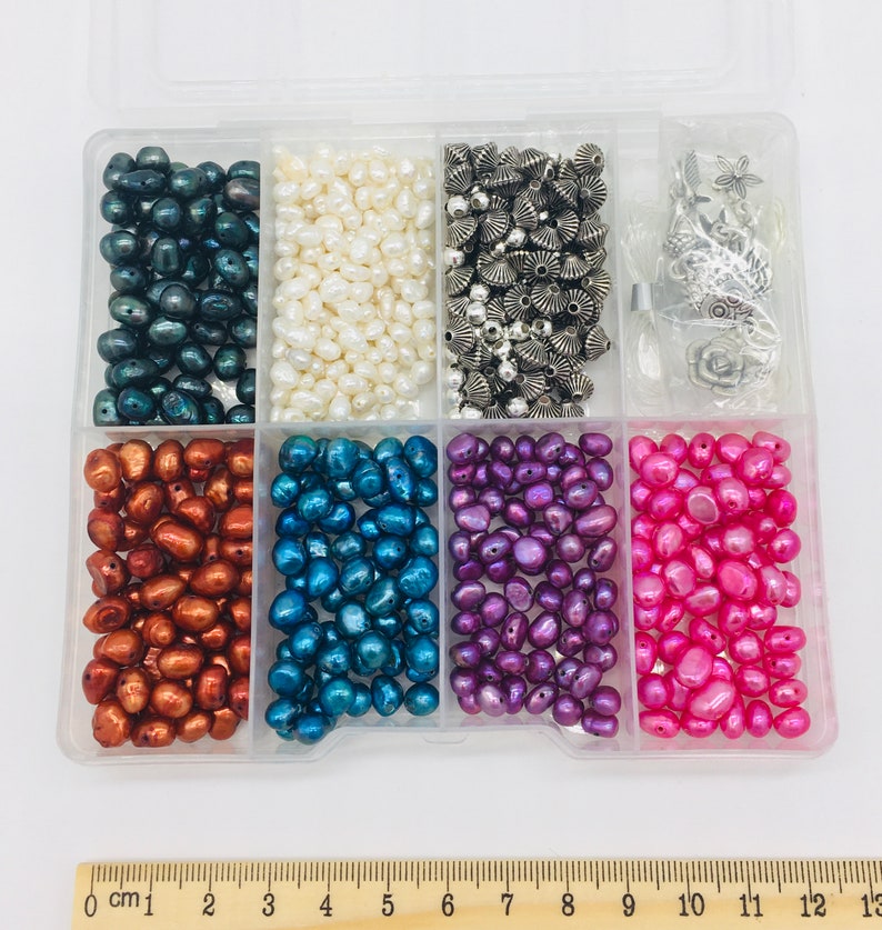 Peut inclure: Une bo&icirc;te de rangement en plastique transparent remplie de diff&eacute;rents types de perles. La bo&icirc;te comporte huit compartiments, chacun rempli d'une couleur de perles diff&eacute;rente. Les perles sont des couleurs suivantes&nbsp;: noir, blanc, argent, cuivre, bleu, violet et rose.