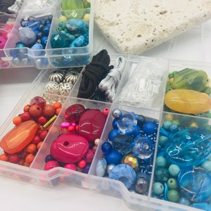 May include: A clear plastic storage box filled with colourful beads, including red, orange, yellow, green, blue, and purple. The box is divided into compartments, and there are also some cords and clasps included.