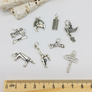 Peut inclure: Une collection de breloques en argent avec diff&eacute;rents motifs, dont une machine &agrave; coudre, un fer &agrave; cheval, un boulier, un bateau, une danseuse, une perceuse et un panneau de camping. Les breloques sont pr&eacute;sent&eacute;es sur une r&egrave;gle en bois avec des marques en centim&egrave;tres.
