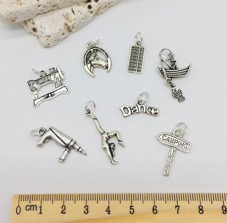 Peut inclure: Une collection de breloques en argent avec diff&eacute;rents motifs, dont une machine &agrave; coudre, un fer &agrave; cheval, un boulier, un bateau, une danseuse, une perceuse et un panneau de camping. Les breloques sont pr&eacute;sent&eacute;es sur une r&egrave;gle en bois avec des marques en centim&egrave;tres.