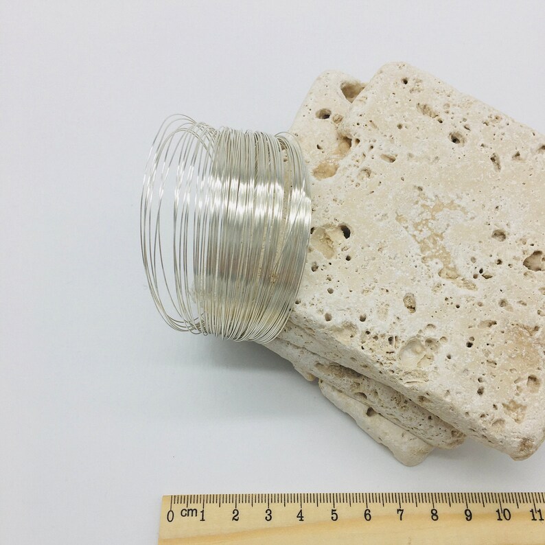 May include: A close-up of a silver wire coil, measuring 11 cm in length, on a beige textured surface. The wire is coiled tightly and appears to be made of thin, flexible wire.
