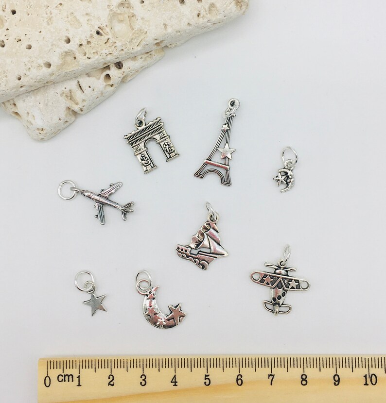 Peut inclure: Un ensemble de neuf breloques en argent avec des motifs sur le th&egrave;me du voyage, notamment un avion, un croissant de lune avec des &eacute;toiles, une &eacute;toile, un voilier, un avion, la tour Eiffel, l'Arc de Triomphe et un croissant de lune.