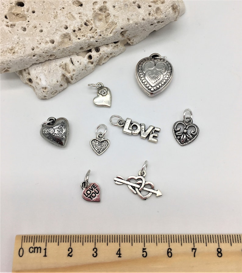 Peut inclure: Assortiment de pendentifs de charme en argent avec des c&oelig;urs, des fleurs et le mot "LOVE". Les breloques sont pr&eacute;sent&eacute;es sur une surface blanche avec une r&egrave;gle en bois pour l'&eacute;chelle.