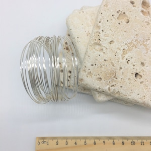 May include: A bundle of 20 silver-colored wire rings, each approximately 1.5 inches in diameter. The rings are stacked on top of each other and are arranged in a circular pattern. A wooden ruler with centimeter markings is in the bottom right corner of the image.