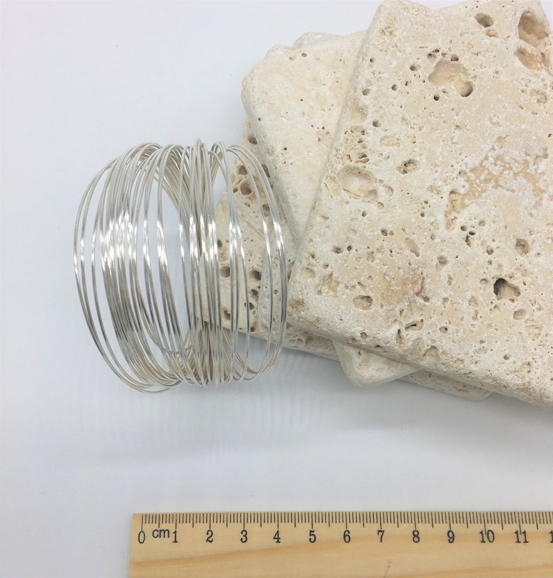 May include: A bundle of 20 silver-colored wire rings, each approximately 1.5 inches in diameter. The rings are stacked on top of each other and are arranged in a circular pattern. A wooden ruler with centimeter markings is in the bottom right corner of the image.