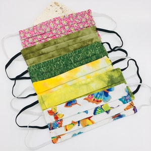 May include: Set of six face masks with different patterns. The masks are made of fabric and have elastic ear loops. The patterns include pink flowers, green grass, yellow tie-dye, and butterflies.