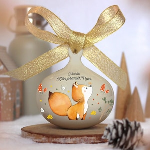 May include: A white ceramic Christmas bauble featuring a fox illustration and the text "Charlie Mon premier Noel". The bauble is decorated with gold glitter and has a gold ribbon.