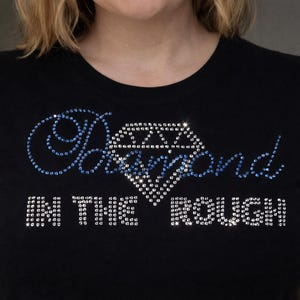 Pode incluir: T-shirt preto com a frase "Diamond in the Rough" em strass. A palavra "Diamond" está em uma fonte azul e a forma do diamante é preenchida com strass.
