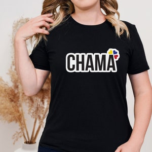Pu&ograve; includere: Una t-shirt nera a maniche corte con la parola CHAMA stampata in grassetto in lettere maiuscole bianche con un contorno nero. A destra del testo si trova un piccolo elemento grafico composto da quattro forme a cuore nei colori giallo, blu e rosso.