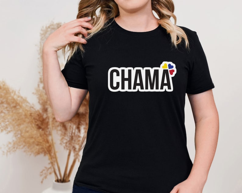 Pu&ograve; includere: Una t-shirt nera a maniche corte con la parola CHAMA stampata in grassetto in lettere maiuscole bianche con un contorno nero. A destra del testo si trova un piccolo elemento grafico composto da quattro forme a cuore nei colori giallo, blu e rosso.