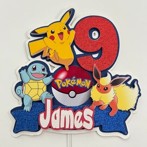 K&ouml;nnte beinhalten: Ein blauer und roter Tortendekor mit einer roten Zahl 9, einem gelben Pikachu, einem blauen Squirtle, einem roten Eevee und einem roten Pok&eacute;ball mit dem Wort "Pok&eacute;mon" darauf. Der Tortendekor hat auch ein blaues Banner mit dem Namen "James" darauf.