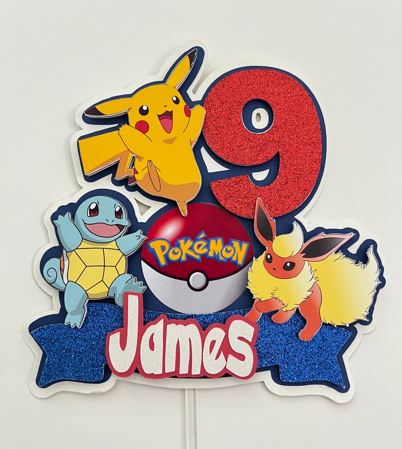 K&ouml;nnte beinhalten: Ein blauer und roter Tortendekor mit einer roten Zahl 9, einem gelben Pikachu, einem blauen Squirtle, einem roten Eevee und einem roten Pok&eacute;ball mit dem Wort "Pok&eacute;mon" darauf. Der Tortendekor hat auch ein blaues Banner mit dem Namen "James" darauf.