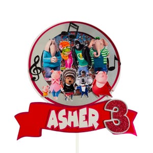 May include: A circular cake topper featuring characters from the movie Sing, including an elephant, a gorilla, and pigs. The center has a shaker element with confetti. A red banner at the bottom displays the name ASHER and the number 3 in glitter.