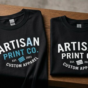 Pu&ograve; includere: Due magliette nere a girocollo con la scritta bianca e azzurra ARTISAN PRINT CO. EST. 2023 CUSTOM APPAREL. La maglietta a sinistra presenta una 'A' azzurra e una grafica azzurra, mentre quella a destra ha testo e grafica di colore bianco.