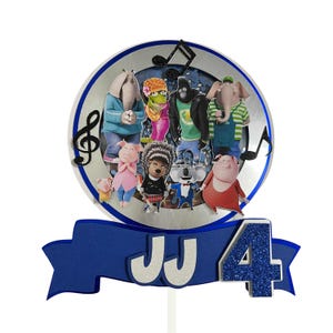 May include: A cake topper featuring characters from the animated movie "Sing." The design includes a silver circle with character cutouts, musical notes, and a blue banner with the letters "JJ" and the number "4" in glitter.