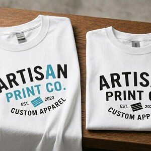 Pu&ograve; includere: Due magliette bianche con il testo nero ARTISAN PRINT CO. CUSTOM APPAREL EST. 2023. La maglietta a sinistra include una A azzurra e una grafica blu, mentre quella a destra presenta testo e grafica interamente di colore nero.