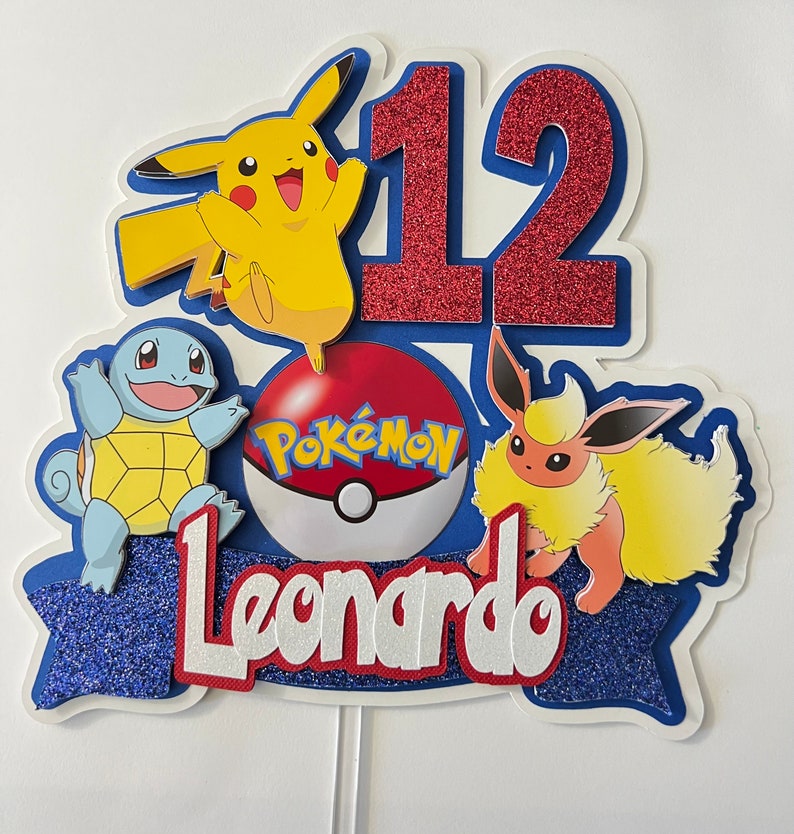 K&ouml;nnte beinhalten: Ein blauer und wei&szlig;er Tortendekor mit einer roten Zahl 12, einem Pok&eacute;ball mit dem Wort Pok&egrave;mon, einem gelben Pikachu, einem blauen Squirtle und einem braunen Eevee. Der Name Leonardo ist in wei&szlig;en Buchstaben auf einem blauen Banner geschrieben.