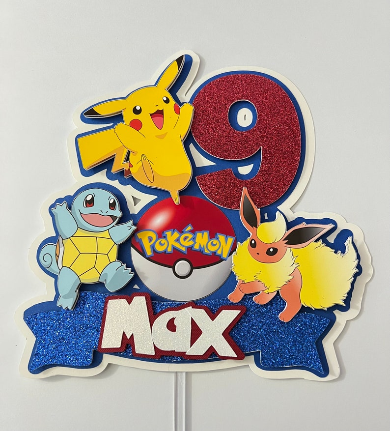 K&ouml;nnte beinhalten: Ein blauer, roter und gelber Pokemon-Kuchentopper mit einer roten Nummer 9, einem gelben Pikachu, einem blauen Squirtle, einem gelben Eevee und einem roten Pokeball mit dem Wort "Pokemon" darauf. Der Topper hat auch ein blaues Banner mit dem Namen "Max" darauf.