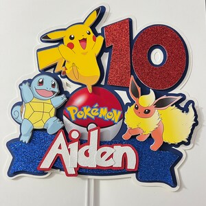 K&ouml;nnte beinhalten: Ein blauer, roter und gelber Kuchentopper zum Thema Pok&eacute;mon mit dem Namen Aiden und der Zahl 10. Der Topper zeigt einen Pok&eacute;ball mit dem Wort Pok&eacute;mon, Pikachu, Eevee und Squirtle.