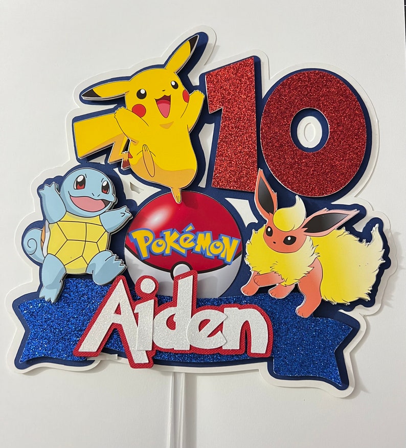 K&ouml;nnte beinhalten: Ein blauer, roter und gelber Kuchentopper zum Thema Pok&eacute;mon mit dem Namen Aiden und der Zahl 10. Der Topper zeigt einen Pok&eacute;ball mit dem Wort Pok&eacute;mon, Pikachu, Eevee und Squirtle.