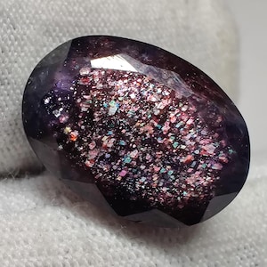 May include: A faceted, oval-shaped gemstone with a dark purple exterior and a sparkling interior. The interior features a mix of pink, blue, and green glitter-like inclusions. The gem is set against a white background.