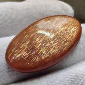 Puede incluir: Gema de piedra solar ovalada y pulida. La piedra tiene un color base marrón rojizo con inclusiones brillantes e iridiscentes que capturan la luz, creando un efecto brillante. La gema es un accesorio natural con tonos tierra. Mide unos 3 cm de largo.