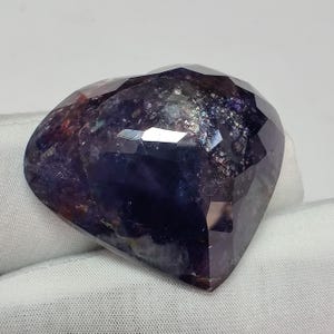 May include: A faceted, heart-shaped gemstone with a deep purple hue, exhibiting a play of colors. The stone's surface reflects light, showcasing a mix of dark blues, purples, and hints of red. The gemstone is set against a white background.